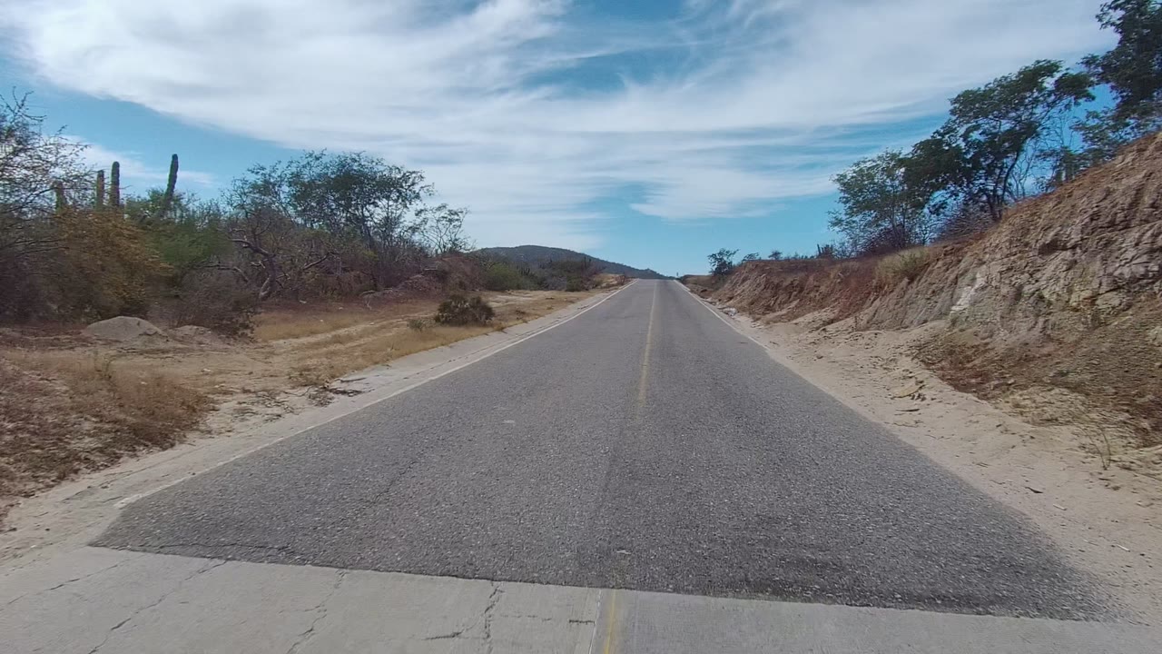 A straight paved road in East Cape Los Cabos, Mexico passes a sandy roadside embankment with brush and cacti-like plants under a partly cloudy blue sky, with no visible people.