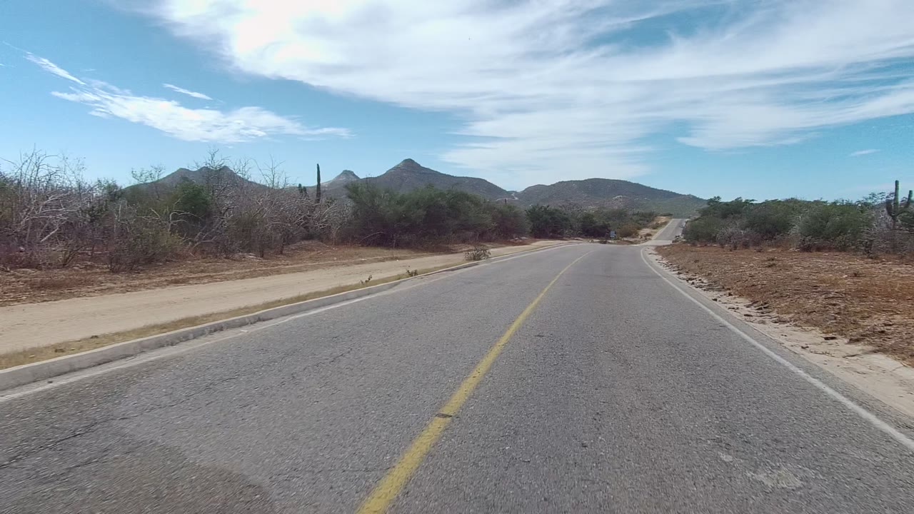 Opening frame of an East Cape Los Cabos paved road with a yellow center line, dry scrub, cacti on the left, low mountains ahead, thin white clouds, and no visible people.