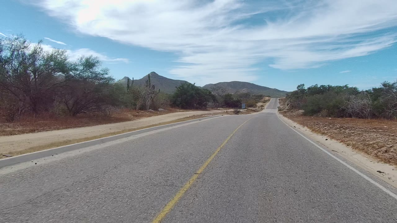East Cape Los Cabos desert road at two seconds, showing the lane centered between sandy shoulders, cactus and brush on both sides, rounded mountains ahead, high blue sky, and no visible people.