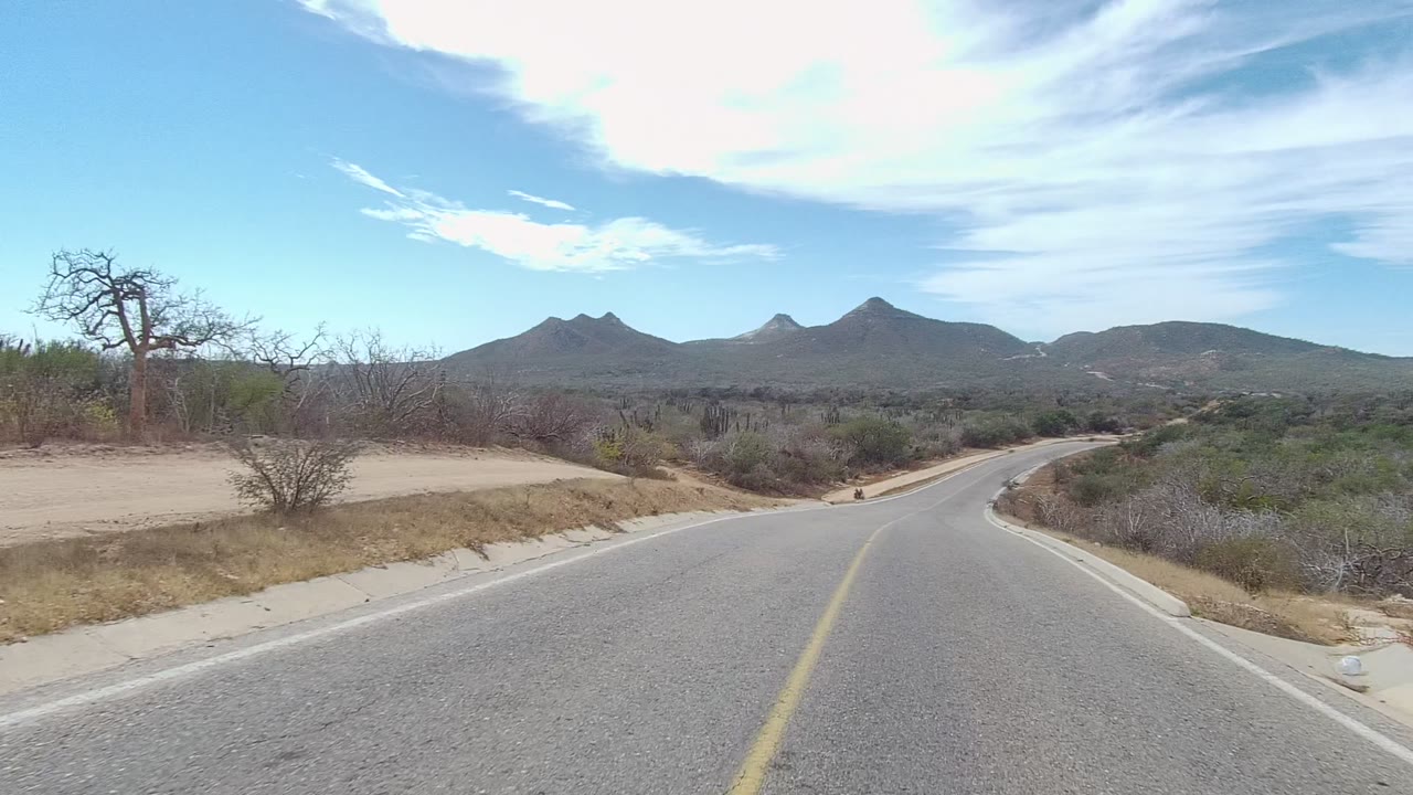 Road-level view in East Cape Los Cabos showing a paved lane, sandy shoulder, desert brush, distant mountains, bright clouds, and no visible people.