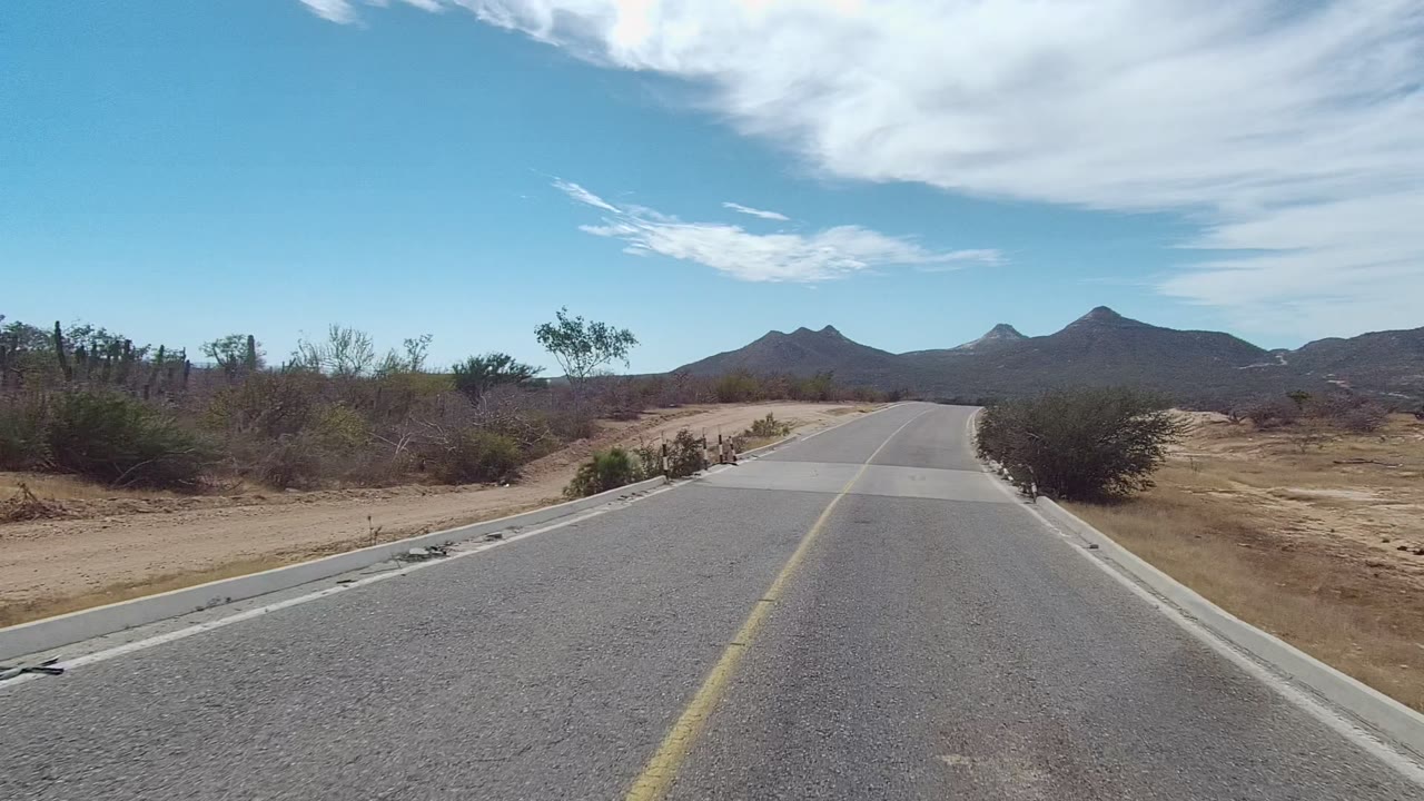Opening frame of a paved road in East Cape Los Cabos, Baja California Sur, with dry roadside brush, distant mountains, broad white clouds, and no visible people.