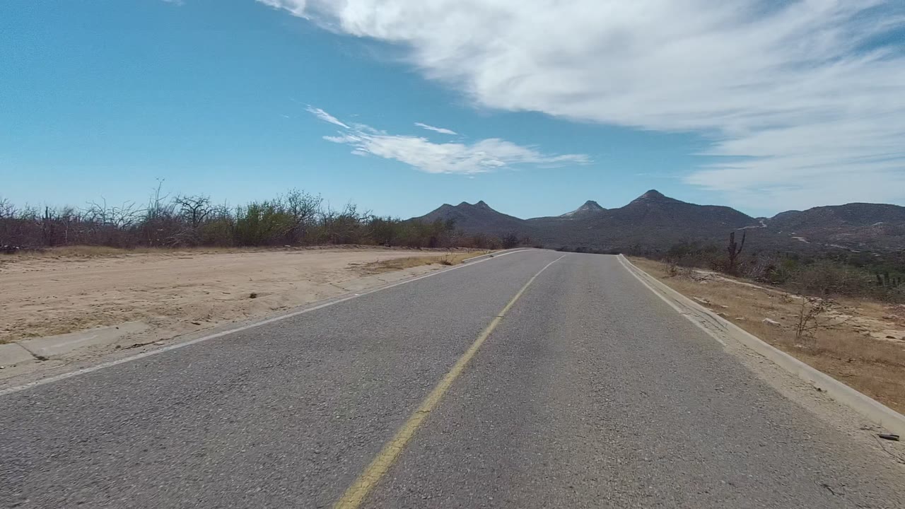 East Cape Los Cabos road view with the asphalt centered, low desert shrubs along both sides, sandy open ground to the right, dark mountain ridges ahead, and no visible people.