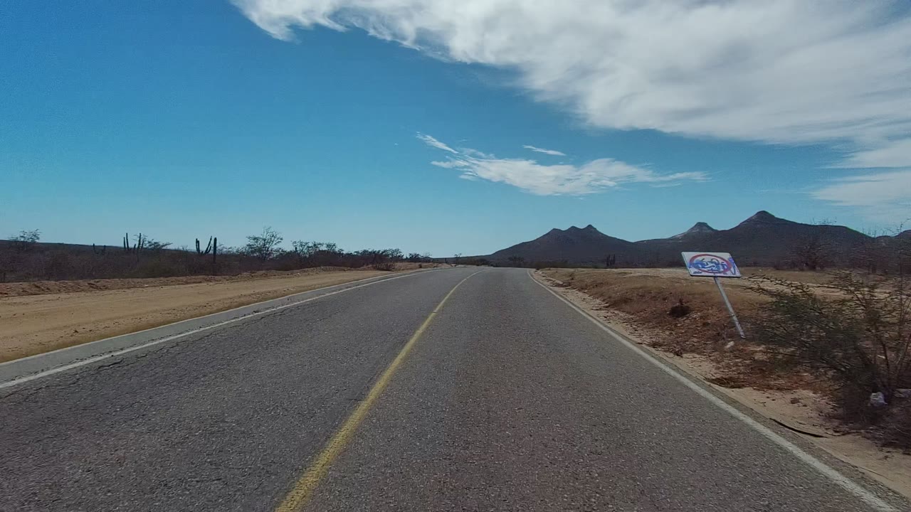 At 0:00 in Los Cabos, Baja California Sur, a paved desert road runs toward distant mountains with sandy shoulders, sparse scrub, a small roadside sign, and no visible people.