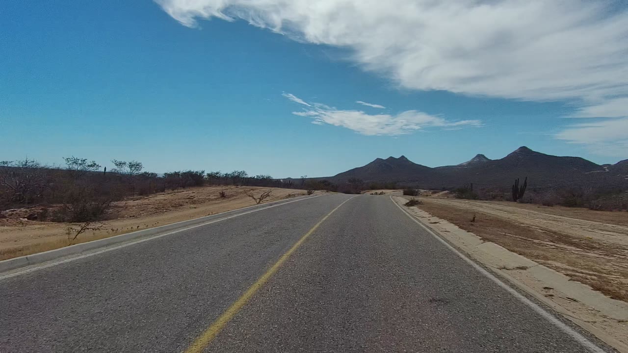 At 0:02 in Los Cabos, Baja California Sur, the highway bends gently through sandy desert terrain with low brush, roadside concrete edges, distant mountains, and no visible people.