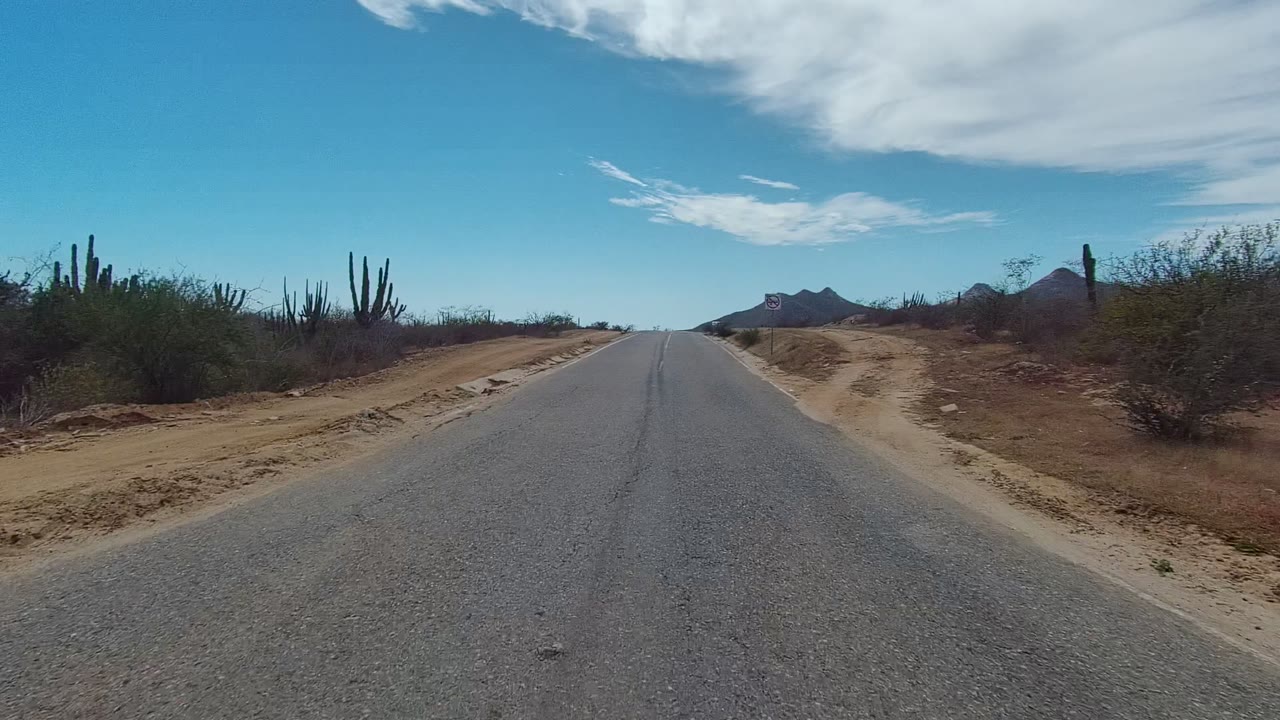 A paved road in East Cape Los Cabos passes a sandy turnout and low desert brush, with tall cacti to the right, distant dark mountains, and no visible people.