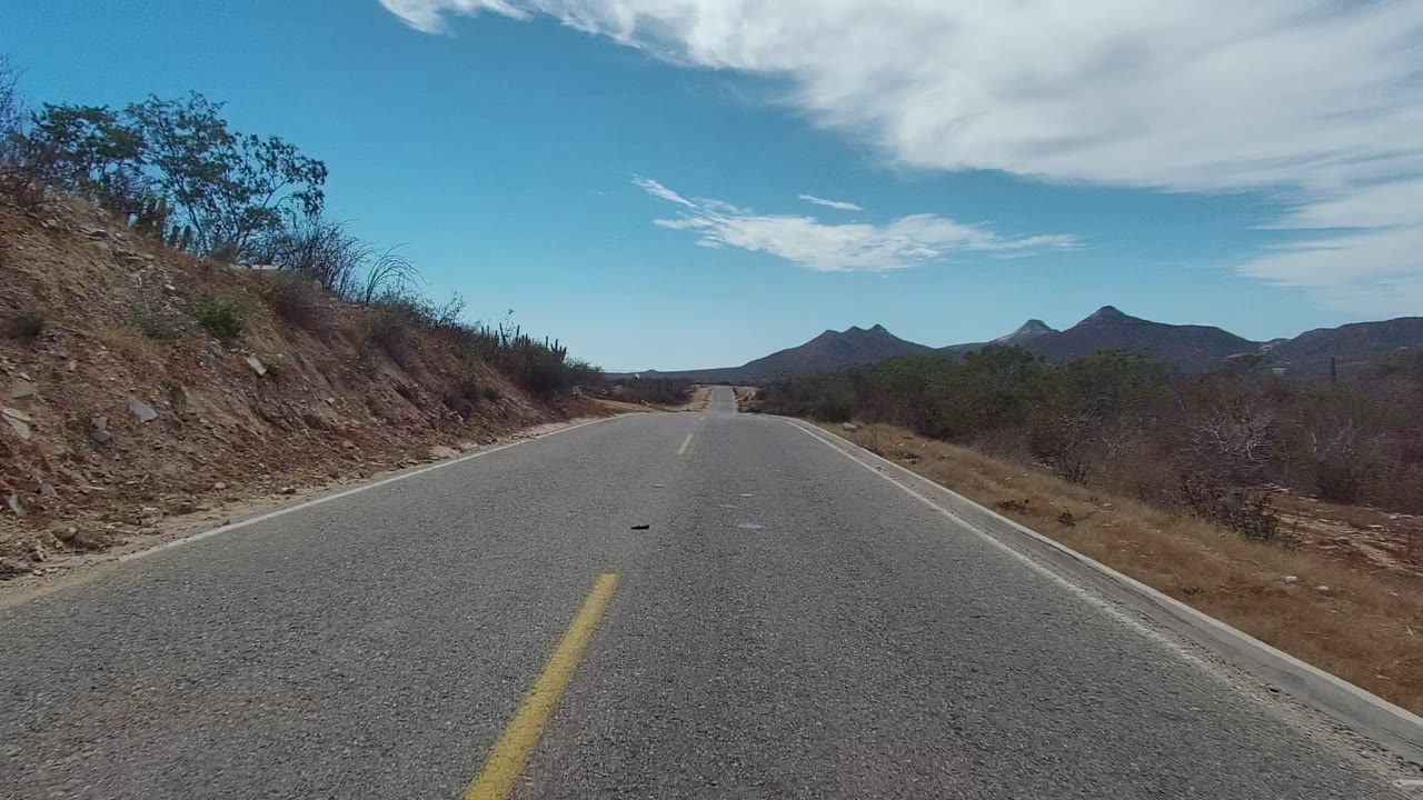 Opening frame of a paved road in East Cape Los Cabos, Baja California Sur, with a yellow center line, rocky left shoulder, dry brush, distant mountains, blue sky, white cloud, and no visible people.