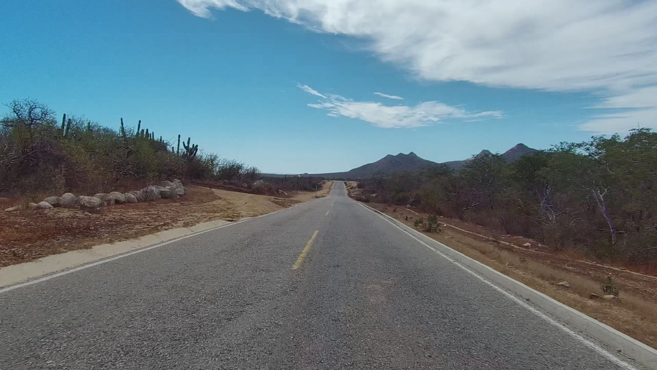 A paved road in East Cape Los Cabos passes a pale dirt side track, roadside rocks, cacti, dry shrubs, distant dark mountains, blue sky, a large white cloud, and no visible people.