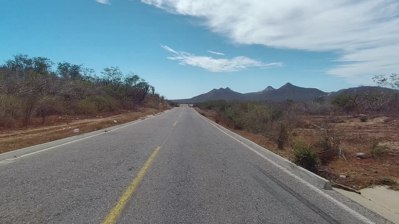 Empty paved road in East Cape Los Cabos crossing dry brushland, with curb edges, low shrubs, distant mountains, and broad white clouds overhead, no visible people.