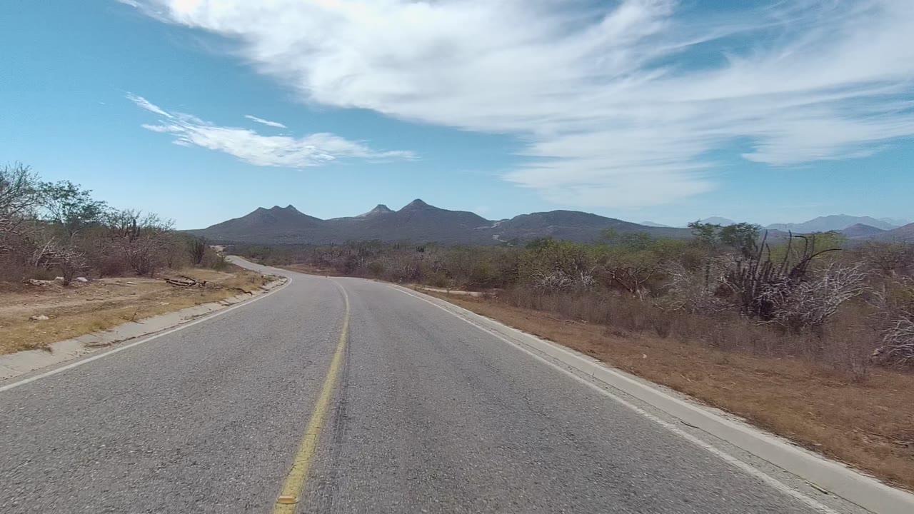 A paved East Cape Los Cabos road fills the foreground as it curves through dry scrub toward dark distant mountains under blue sky, with no visible people.