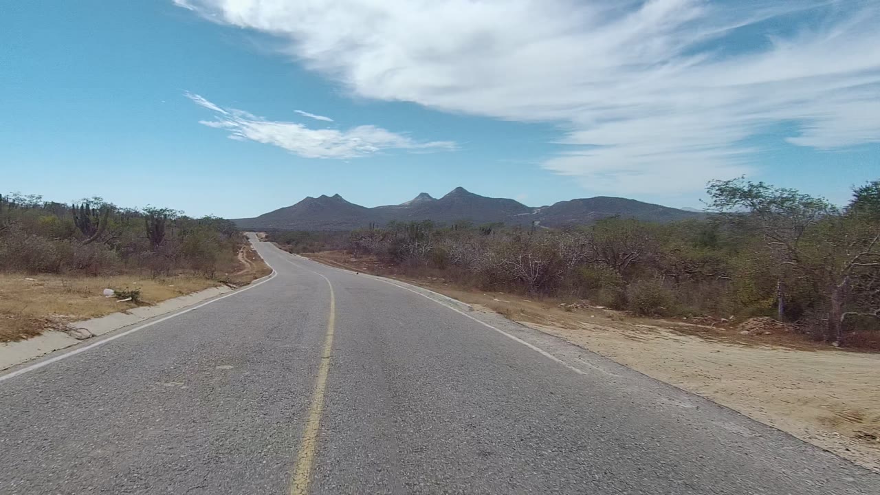 An empty paved road in East Cape Los Cabos passes sparse desert vegetation, with a pale shoulder, distant mountains, broad white clouds, and no visible people.