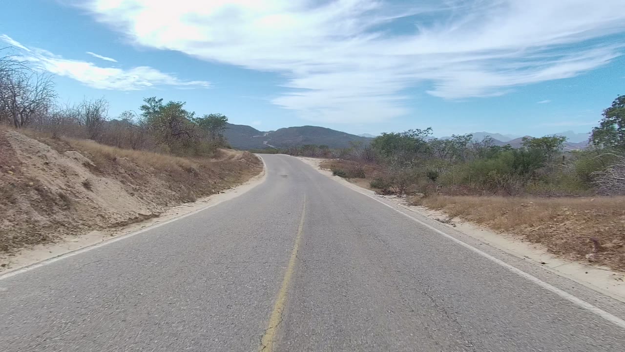 At 0:00, a paved road in East Cape Los Cabos curves ahead between sandy shoulders and dry roadside brush, with distant mountains under a blue sky and no visible people.