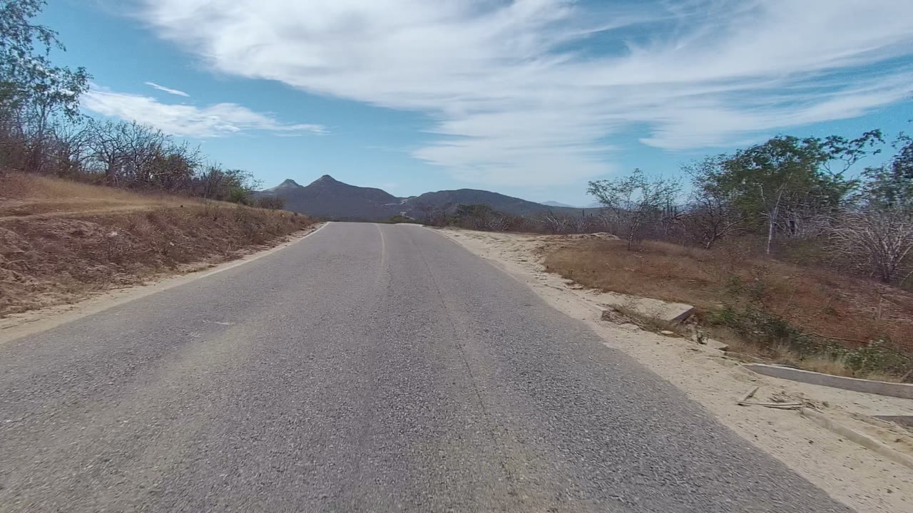 At 0:02, the road runs straight ahead through East Cape Los Cabos, with a dry embankment on the left, sparse brush on both sides, dark mountain peaks, and no visible people.