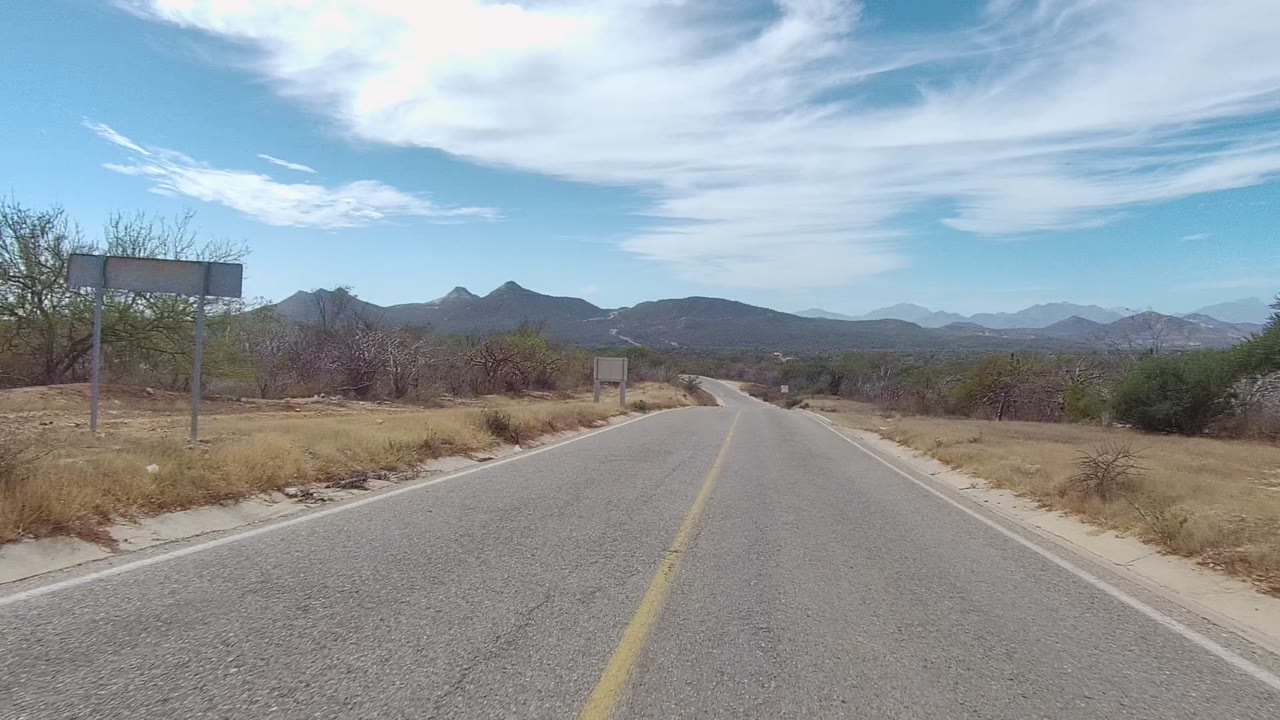 Opening frame of an East Cape Los Cabos road with a yellow center line, dry roadside grass, sparse trees, distant mountains, streaked white clouds, and no visible people.