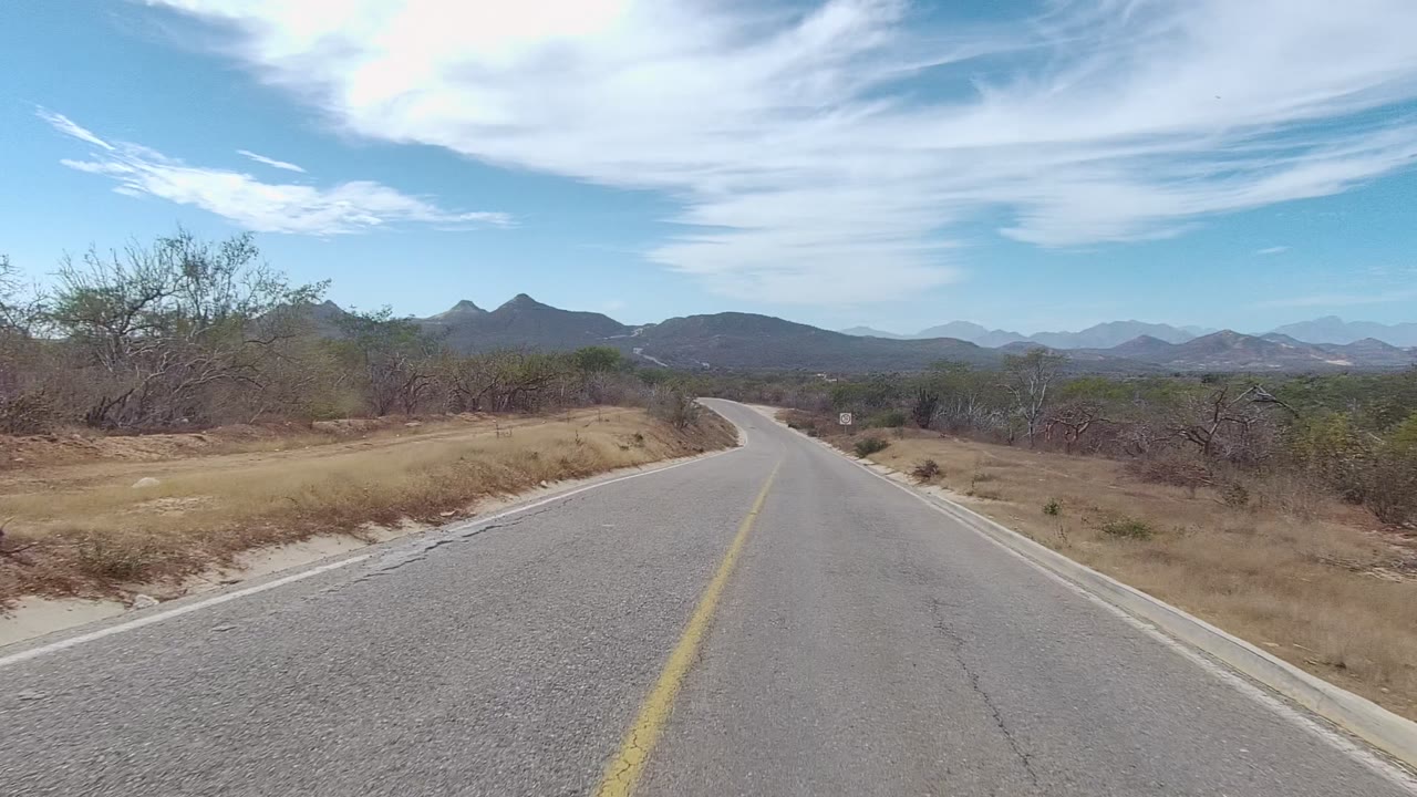 A paved East Cape Los Cabos road begins to bend right through dry grass and low trees, with layered mountains in the distance and no visible people.