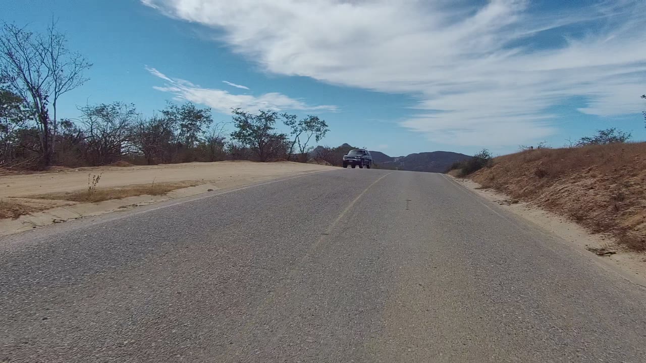 Black SUV approaches on a paved road in East Cape Los Cabos, Baja California Sur, with dry brush, sandy shoulders, distant mountains, blue sky, and no visible people.