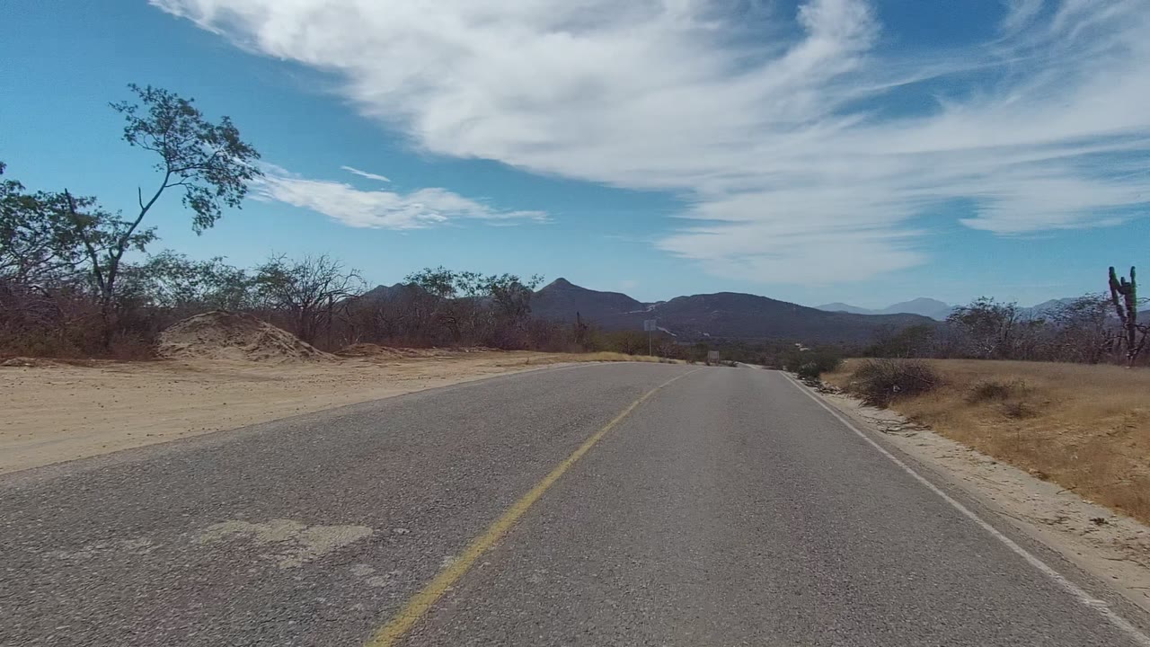 The SUV has left the frame, leaving an empty paved road in East Cape Los Cabos with a yellow line, sandy roadside, cactus, dry scrub, mountains, and no visible people.
