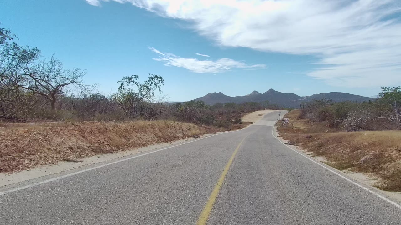 Rural asphalt road in East Cape Los Cabos, Baja California Sur, with a yellow center line, dry grass shoulders, sparse trees, low hills, bright clouds, and no visible people.