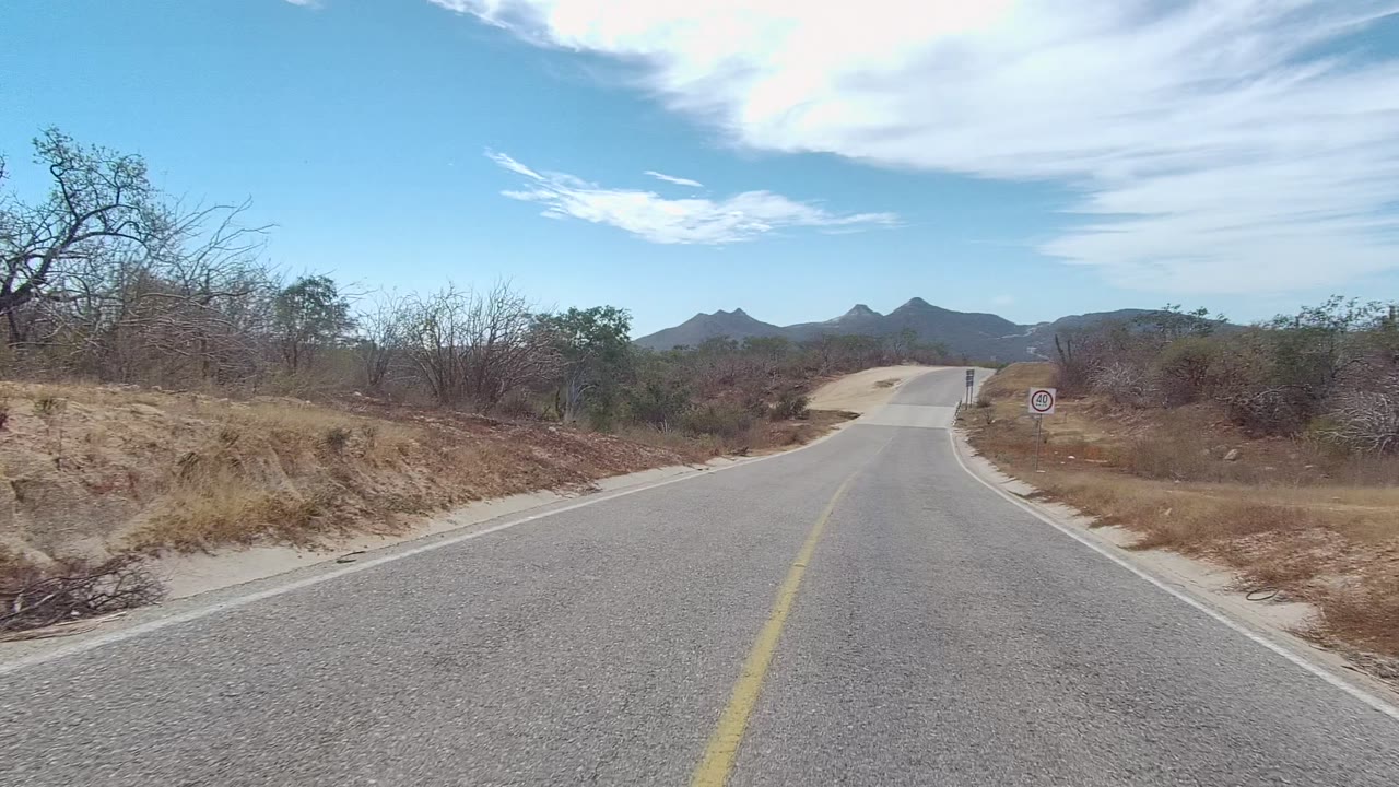 Empty road ahead with dry roadside brush and low hills in the distance.