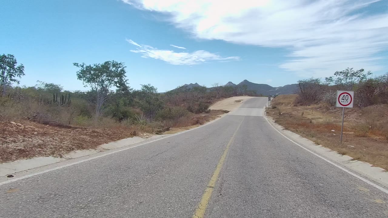Rural road in East Cape Los Cabos, Baja California Sur, with a visible 40 speed limit sign on the right shoulder, scrub vegetation, dry ground, distant hills, and no visible people.