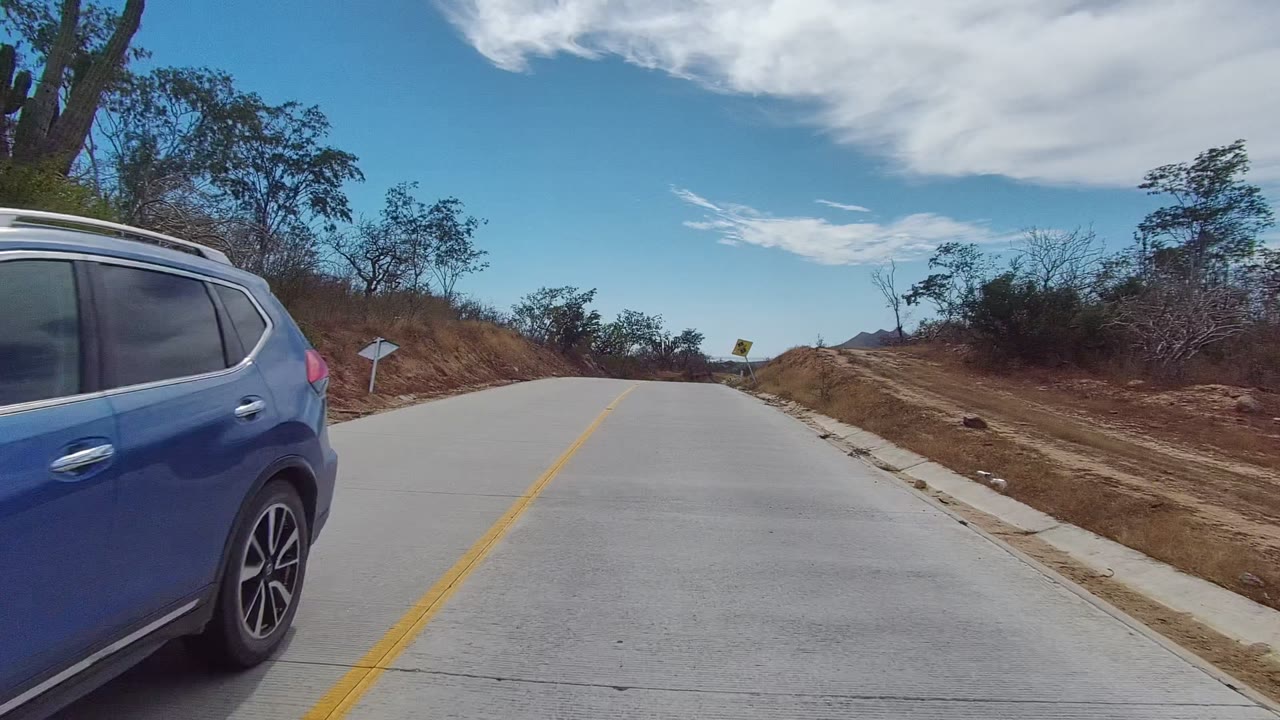 At the start of the Los Cabos clip, a blue car is close on the left edge beside a paved road with a yellow line, dry brush, scattered trees, distant hills, and no visible people.
