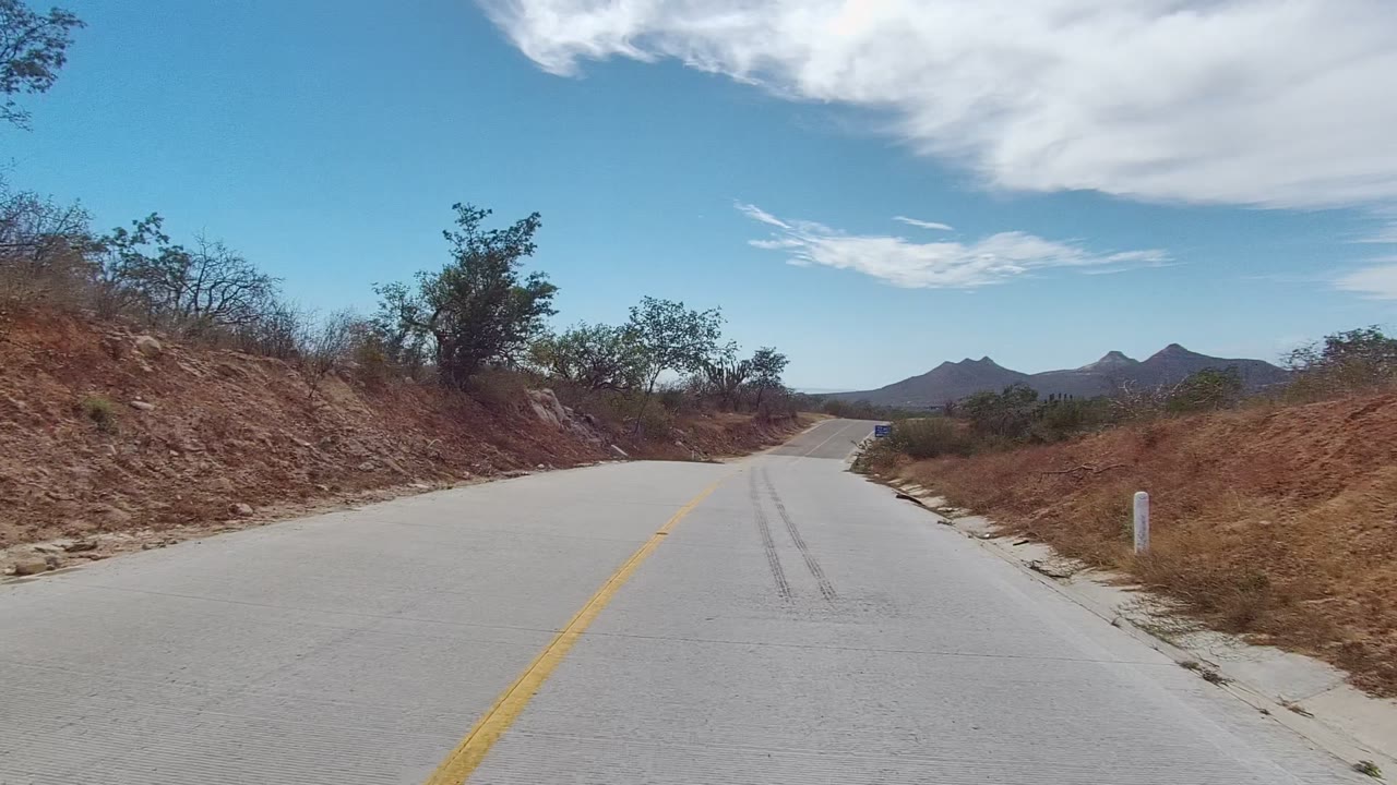 At two seconds, the paved Los Cabos road runs forward past dry brush and low trees toward distant mountains, with a broad blue sky, white clouds, and no visible people.