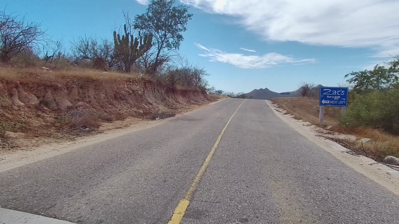 A paved two-lane road in East Cape Los Cabos, Baja California Sur, with dry banks, cactus, a blue roadside sign, distant mountains, and no visible people.