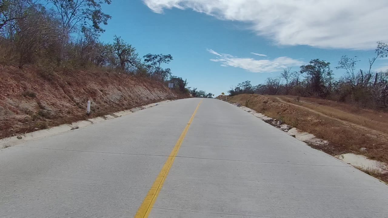 A pale paved road in East Cape Los Cabos passes reddish dirt banks and sparse trees, with no visible people in the frame.