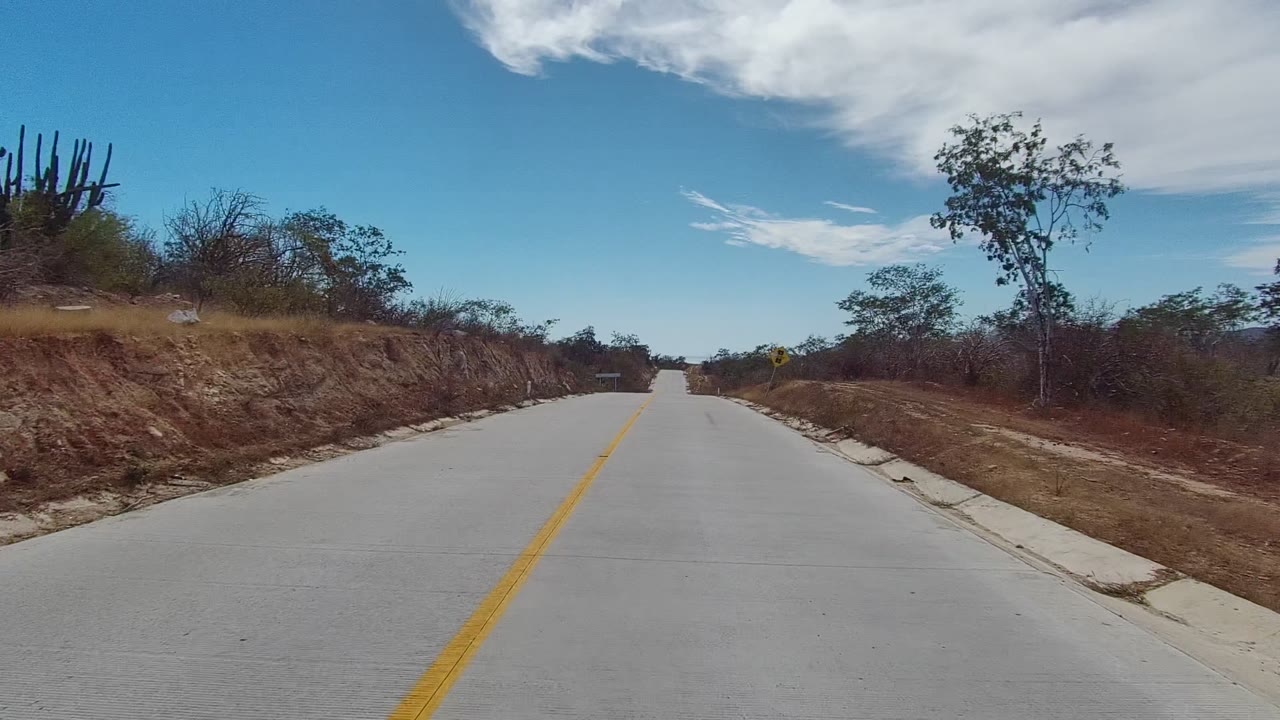 Forward view of a paved East Cape Los Cabos road with a yellow center line, dry brush, cactus at the left edge, blue sky, white clouds, and no visible people.