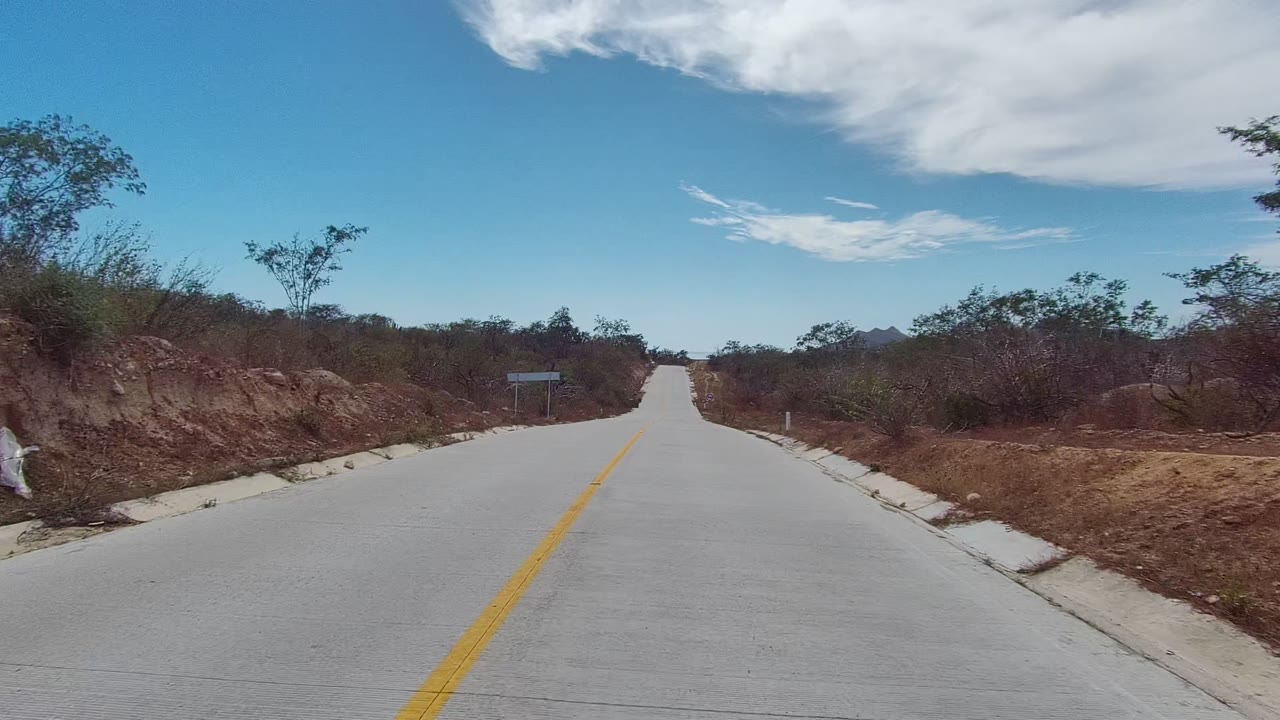 A straight paved road in East Cape Los Cabos runs between reddish dirt shoulders, sparse desert shrubs, small signposts, bright sky, and no visible people.