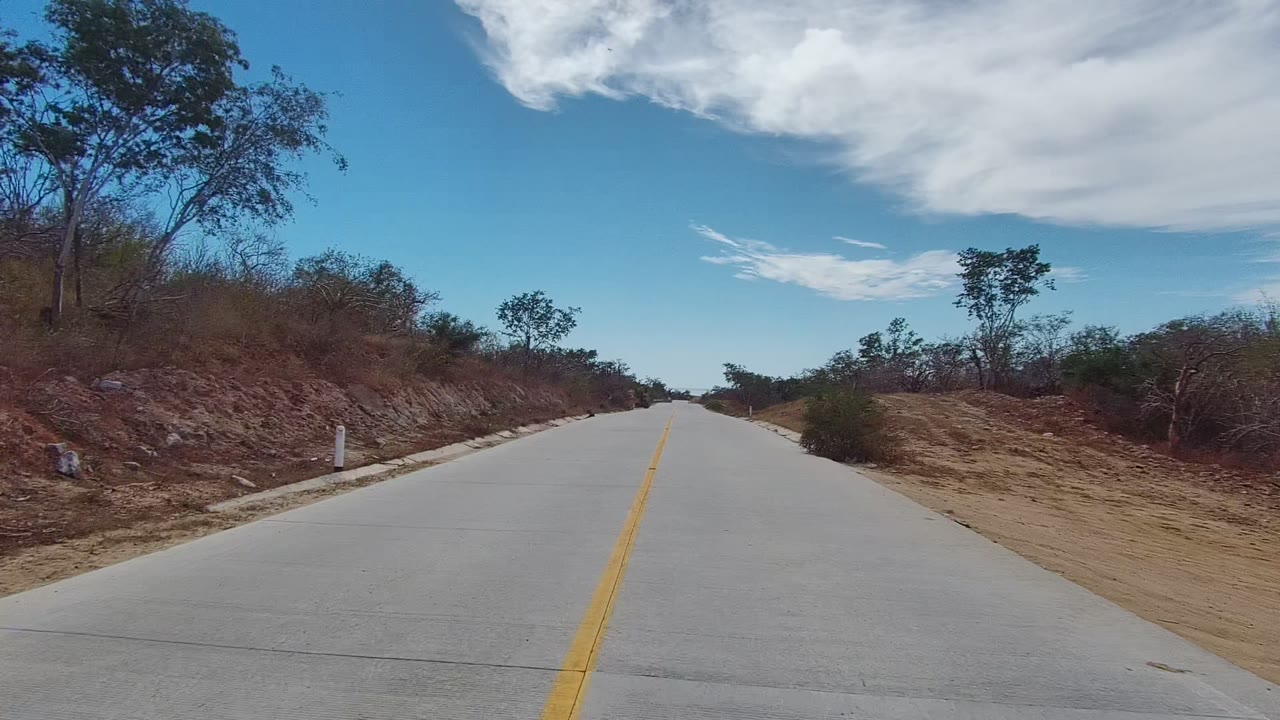 A paved road in East Cape Los Cabos, Baja California Sur runs toward the horizon between dry brush, small trees, and a blue sky with white clouds, with no visible people.