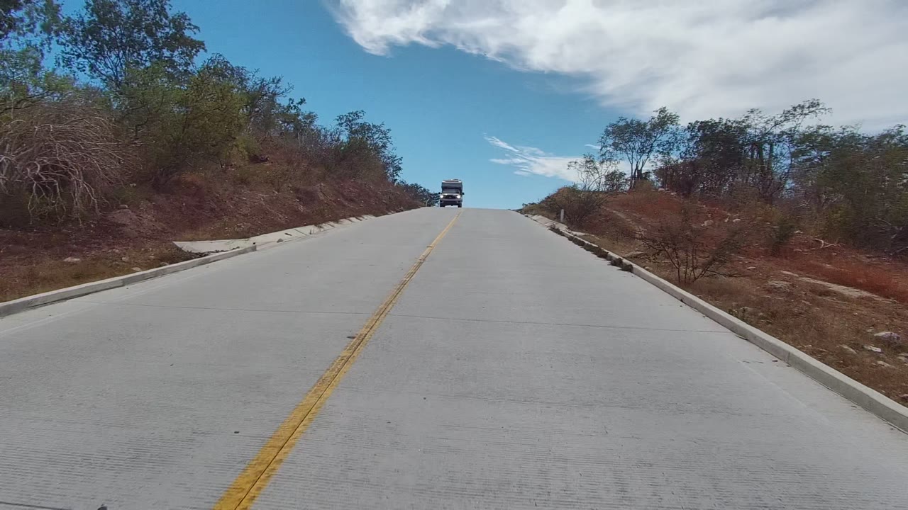A distant truck approaches on a pale concrete road in East Cape Los Cabos, Mexico, with dry roadside vegetation, blue sky, white clouds, and no visible people.