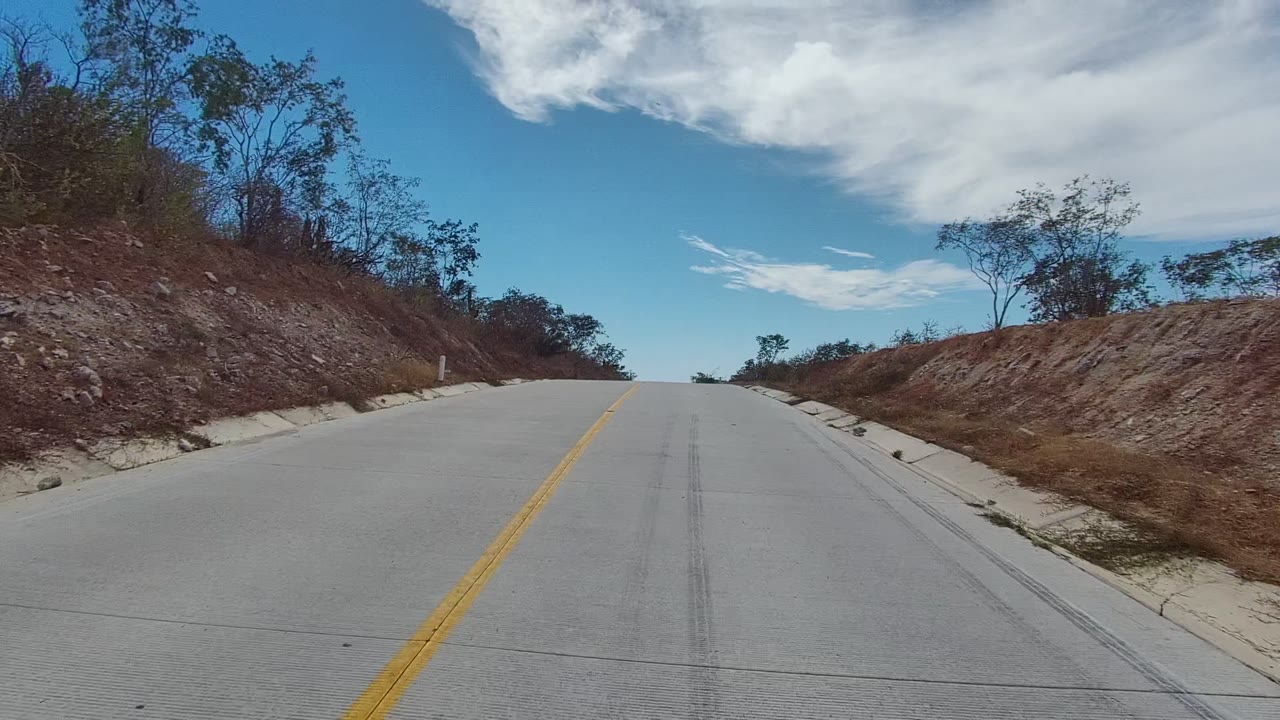The truck has passed out of view, leaving an empty uphill concrete road in East Cape Los Cabos, Mexico, bordered by dry slopes, scattered trees, white clouds, and no visible people.