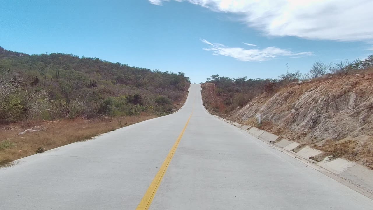 At 0:00, a paved road in East Cape Los Cabos, Baja California Sur climbs straight ahead between dry brushy hillsides, with a yellow center line and no visible people.