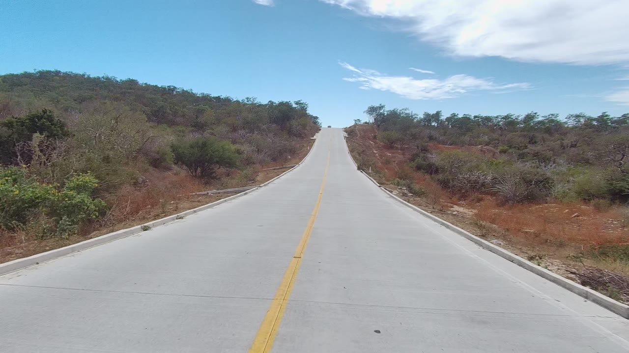 At 0:02, an empty paved road in East Cape Los Cabos runs toward a hill crest between scrub-covered brown slopes, with no visible people.