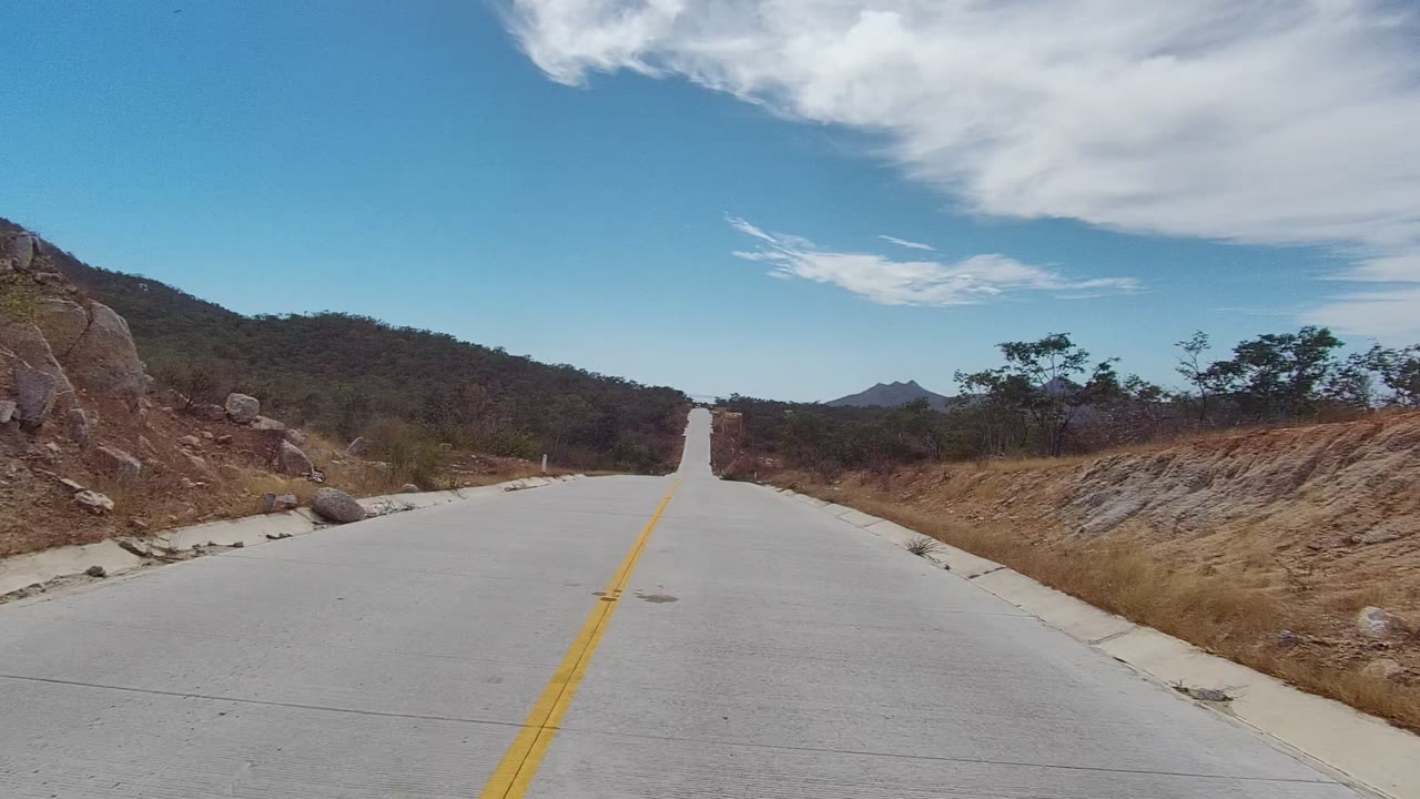 At the start of the East Cape Los Cabos clip, a pale paved road with a yellow center line leads into dry hills and scrub beneath a blue sky, with no visible people.