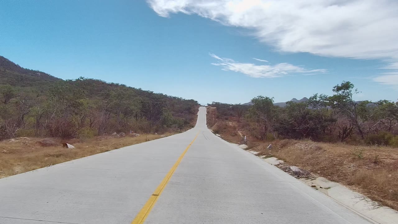 Two seconds in, the road stretches ahead through East Cape Los Cabos scrubland, with a hillside on the left, trees on both sides, and high white clouds, with no visible people.