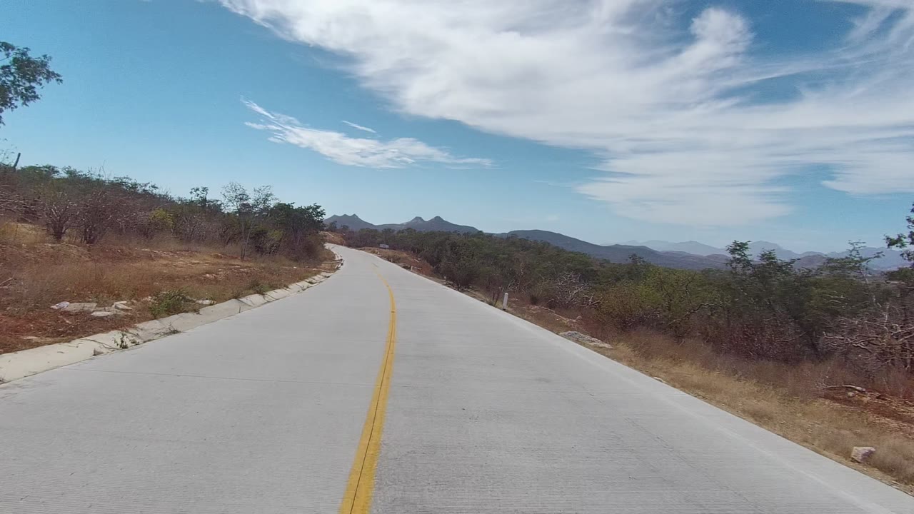 A paved East Cape Los Cabos road curves ahead between dry brush and scattered trees, with distant mountains and broad white clouds overhead, no visible people.