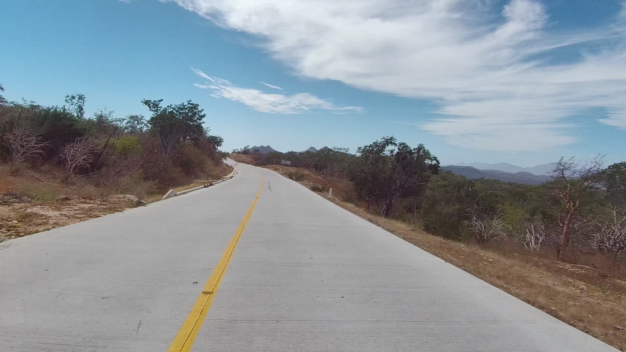 A later view down the East Cape Los Cabos road shows the right bend more clearly, with brush close to the shoulders, mountains on the horizon, and high white clouds, no visible people.