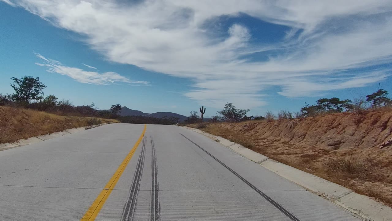 Opening frame of an East Cape Los Cabos road with skid marks on pale pavement, dry roadside brush, a cactus, distant mountains, white clouds, and no visible people.
