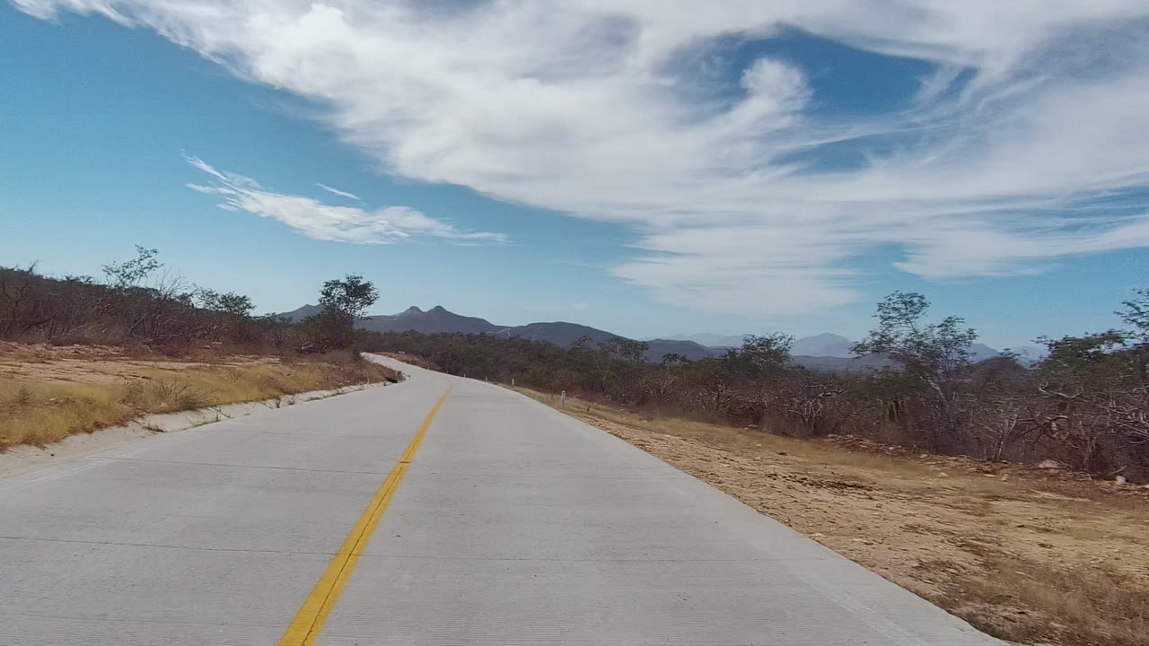 The road continues through East Cape Los Cabos scrubland, with the yellow line leading into a right curve, low hills ahead, dry shoulders, layered clouds, and no visible people.