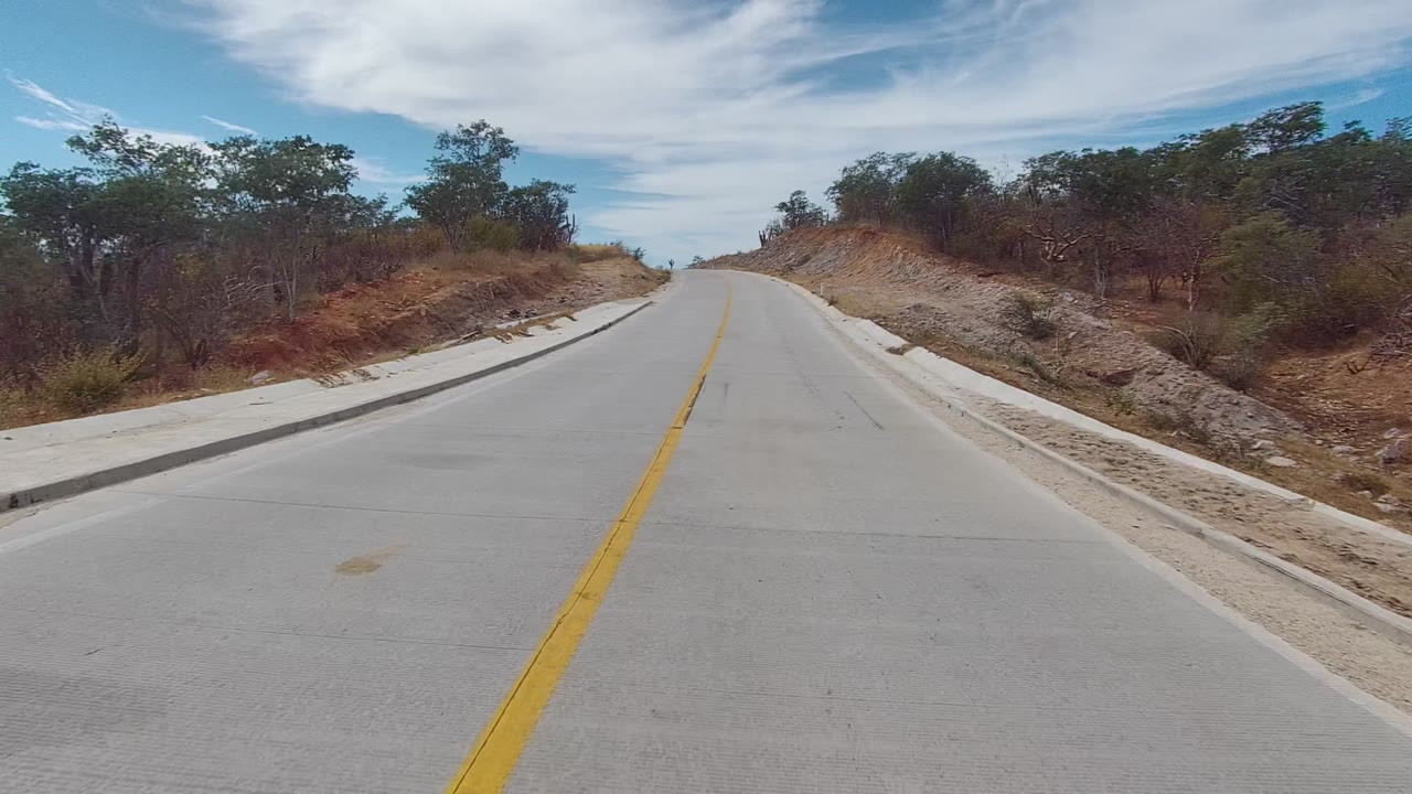 A paved road in East Cape Los Cabos, Baja California Sur rises between dry roadside banks, sparse trees, and white clouds, with no visible people.