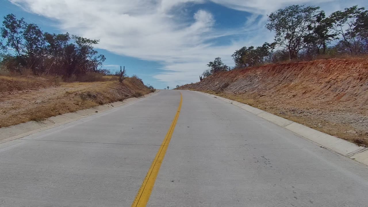 A forward road view in East Cape Los Cabos shows the pavement bending slightly between red-brown cut banks, scattered trees, cactus shapes, and no visible people.