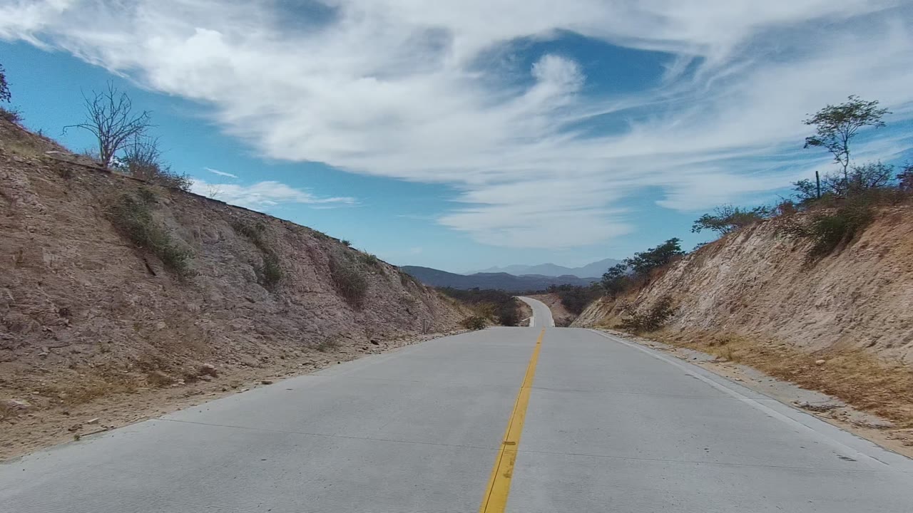 A paved road in East Cape Los Cabos, Baja California Sur curves between pale rocky hillsides, sparse shrubs, distant mountains, and broad white clouds, with no visible people.