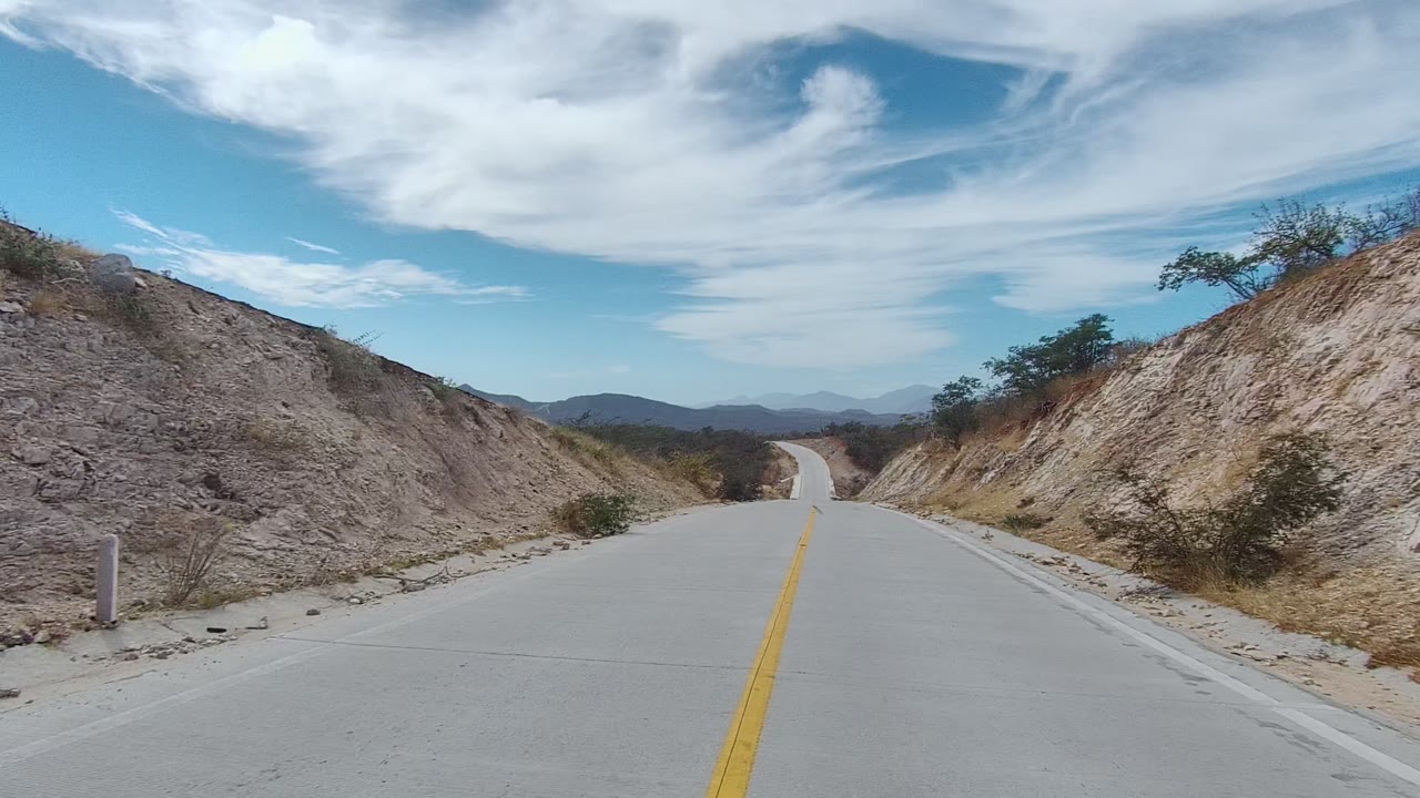 Yellow-lined road running past a pale hillside with mountains ahead.