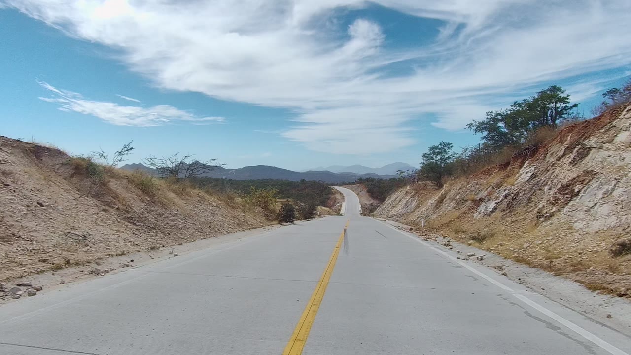 An empty paved road in East Cape Los Cabos approaches a slight bend between dry roadside banks, scattered bushes, low mountains, and a bright blue sky with long cloud bands, with no visible people.