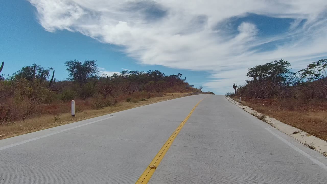 East Cape Los Cabos paved road rises ahead between dry roadside vegetation and cacti beneath wide white clouds, with no visible people.