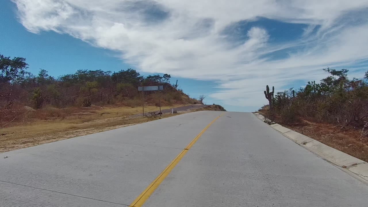 East Cape Los Cabos road approaches a low crest with cactus and brush on both sides and a small roadside sign structure visible at right, with no visible people.