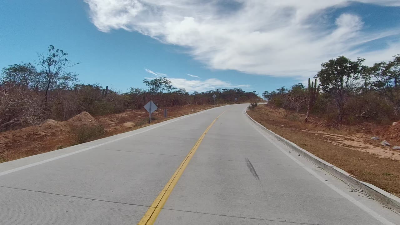 Start of a vehicle-height view on a two-lane road in East Cape Los Cabos, Mexico, with yellow center lines, sparse dry trees, a roadside sign, and no visible people.