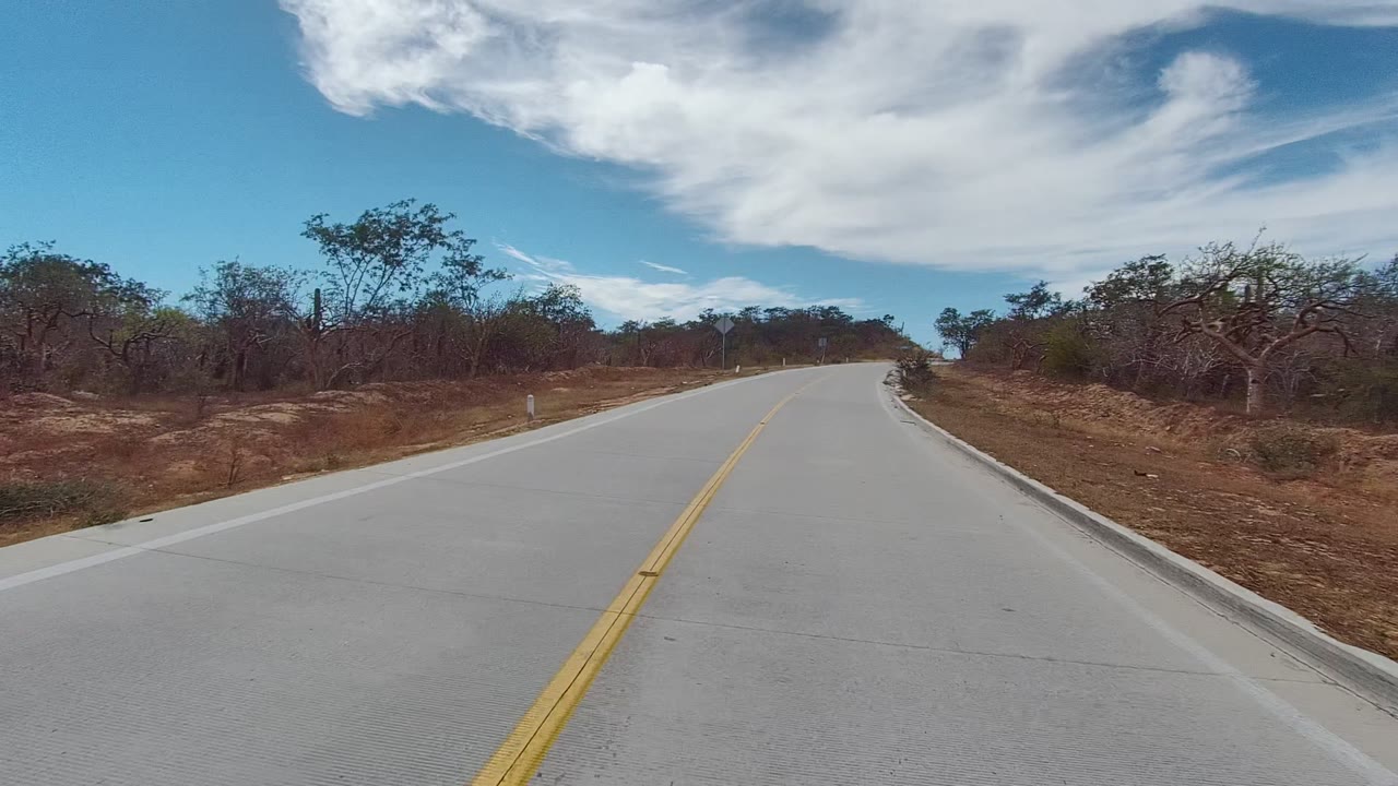 Straight view down a pale two-lane road bordered by brown grass and scrub.