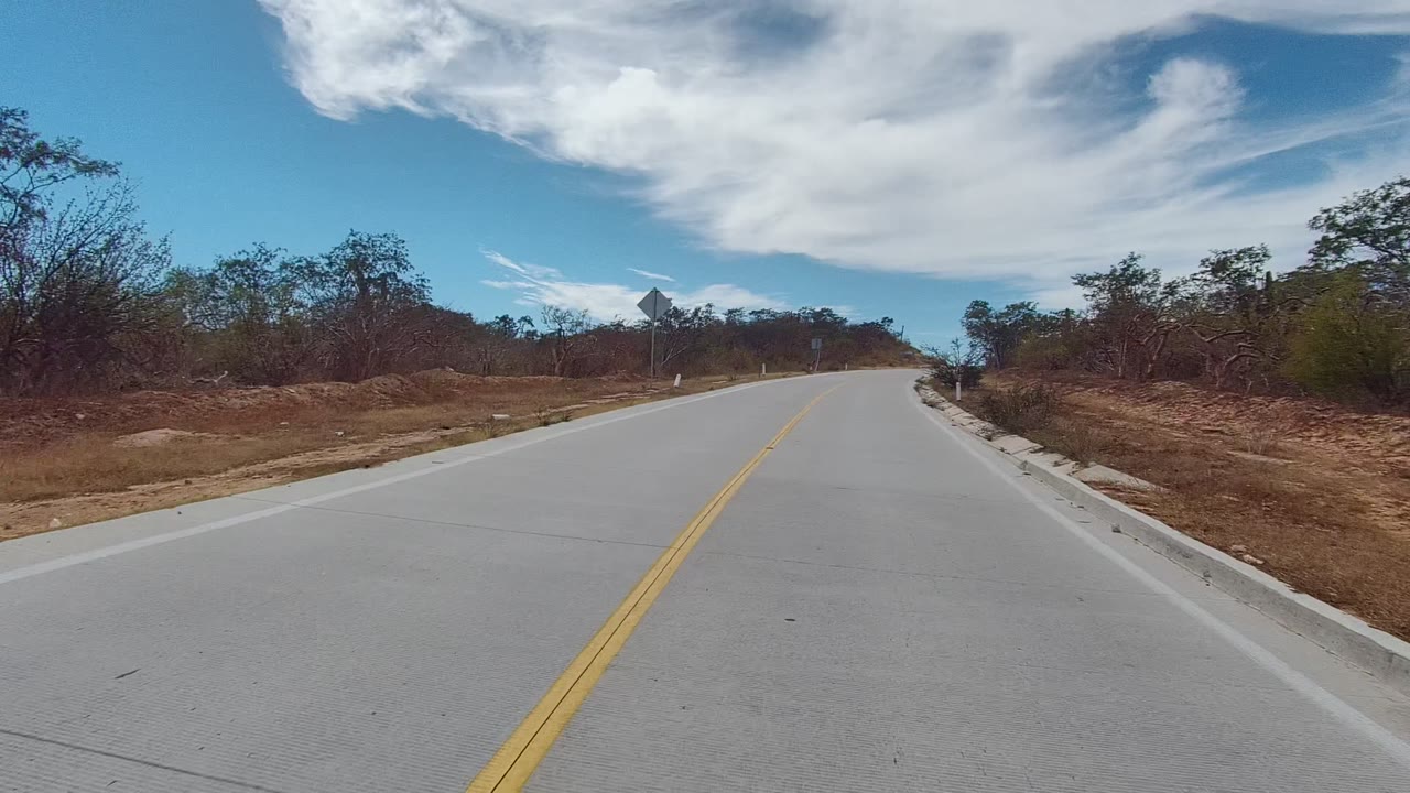 The road continues through East Cape Los Cabos, Mexico, with the yellow lines leading toward a slight rise, dry scrub on both shoulders, a small sign, and no visible people.